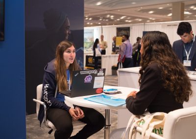Two people sitting at a table in a conference setting, one using a laptop and the other listening attentively. Various booths and attendees are visible in the background.