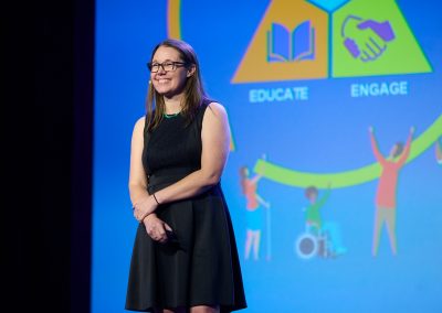 A woman stands on stage in front of a screen displaying visuals with the words "Educate" and "Engage.