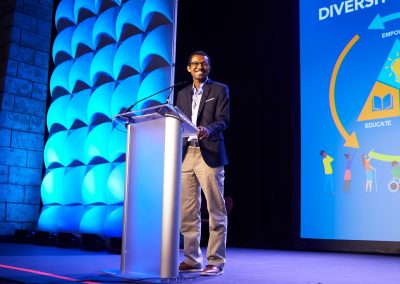 A man in a blazer stands at a podium on a stage, speaking. A presentation slide with bright graphics is displayed on the screen beside him.