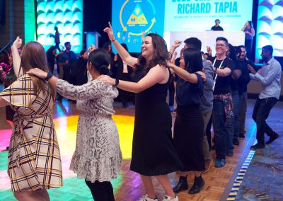 People in a conga line are dancing in a colorful, well-lit event space. A stage with a DJ is visible in the background.