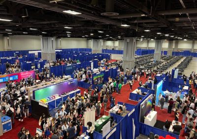 Bird's-eye view of a bustling convention center hall with numerous exhibition booths and attendees interacting, surrounded by blue partitions.