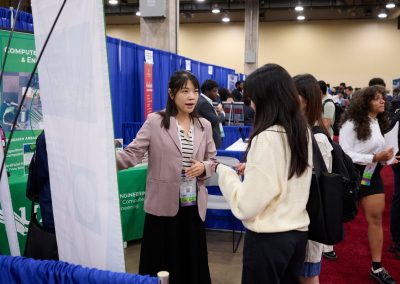 A woman in a blazer speaks with three people at a booth during a conference. The booth displays information about computer engineering.