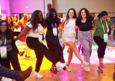 A group of people dance in a circle on a lit-up floor, smiling and holding hands.