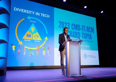 A person stands at a podium on stage during the 2023 CMD-IT/ACM Richard Tapia Conference, with a presentation slide on diversity in tech displaying behind them.