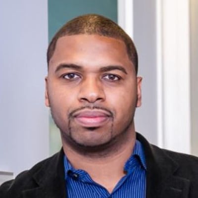 A person with short-cropped hair and a goatee, wearing a blue shirt and dark jacket, is standing indoors at the 2024 conference among notable plenary speakers.