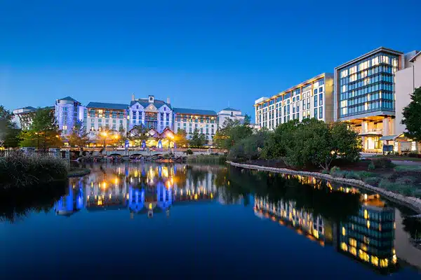At dusk, the large building complex, a renowned venue for Tapia, displays its lit windows reflecting on a serene pond. Framed by trees and shrubs, the scene unfolds under a clear blue sky.