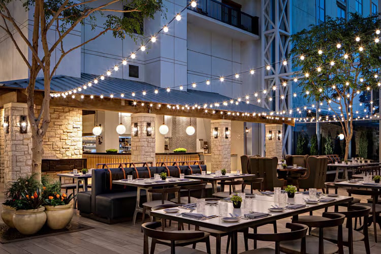 The outdoor restaurant seating area, perfect as a venue for tapia, features tables and chairs beneath charming string lights. Stone walls and large potted plants beautifully encircle the inviting space.