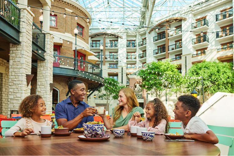A family of five sits at a table in a spacious, well-lit indoor restaurant, the ideal venue for Tapia. They are smiling and enjoying a meal together amidst lush greenery and the charm of a multi-story building.