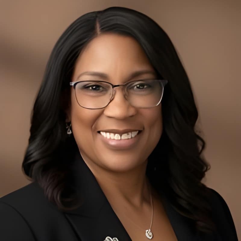 A woman with long dark hair, wearing glasses, a black blazer, and a necklace, smiling in front of a brown background.