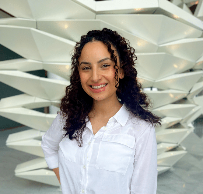 A woman with curly hair wearing a white shirt stands smiling in front of a geometric white architectural structure.
