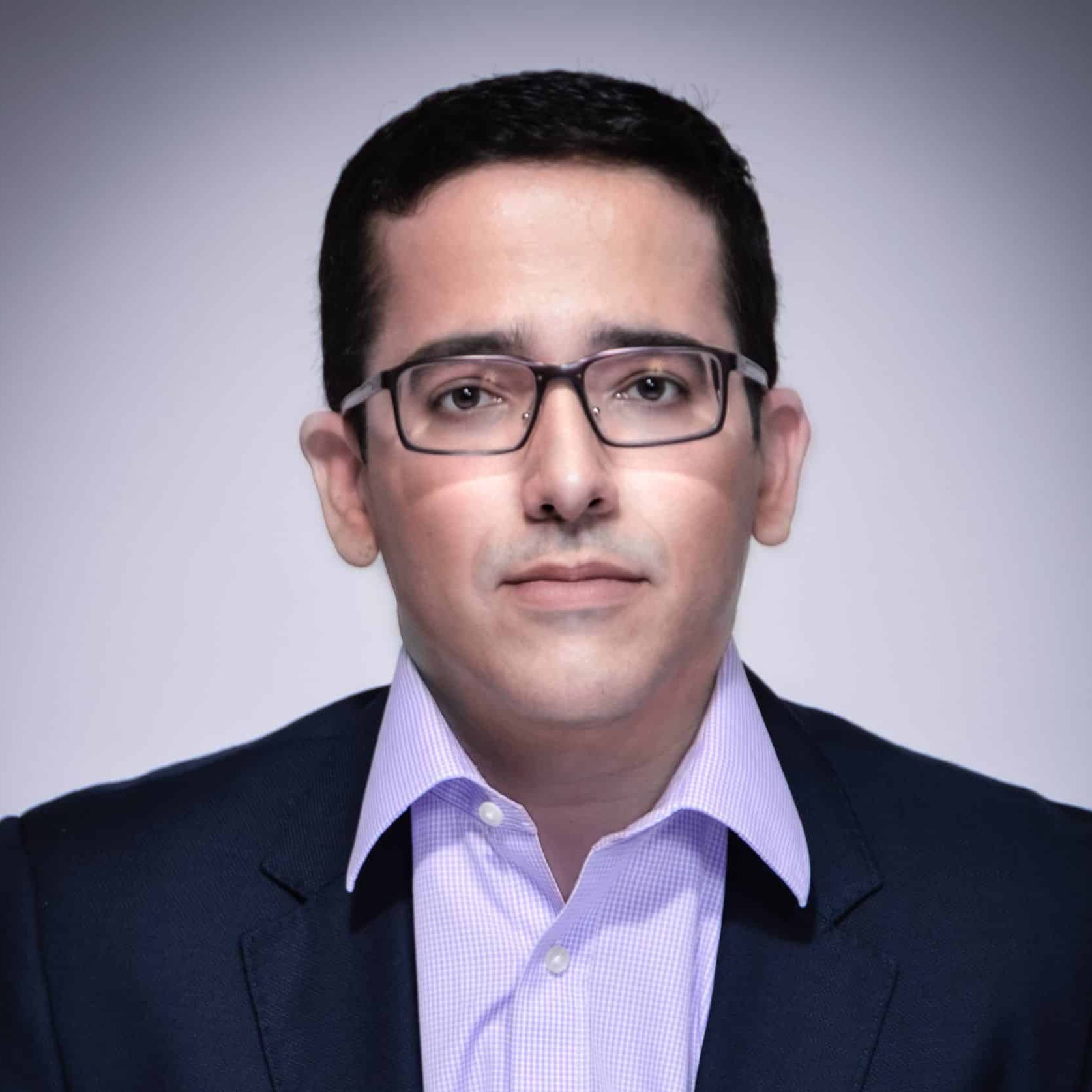 A man wearing glasses and a dark suit over a light purple collared shirt stands against a plain, gradient background, looking directly at the camera.