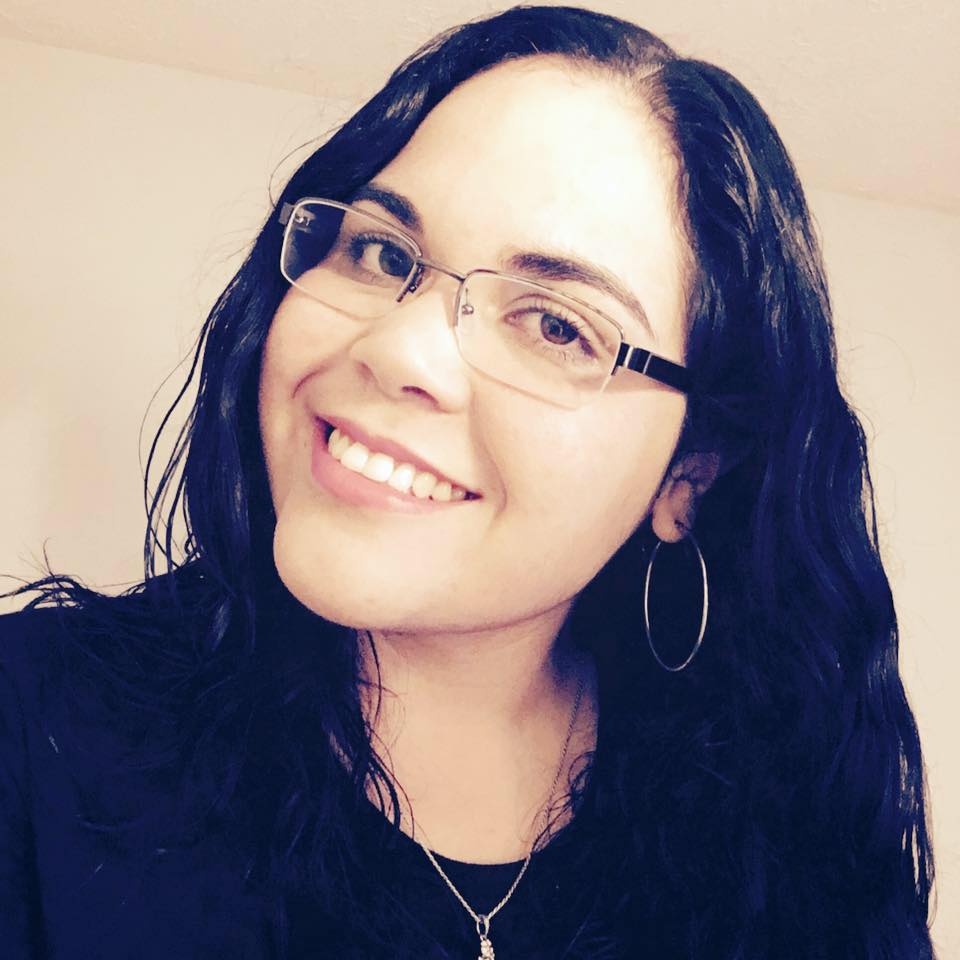 Woman with long dark hair, glasses, and hoop earrings smiles at the camera, wearing a black top and a necklace, against a plain light-colored background.