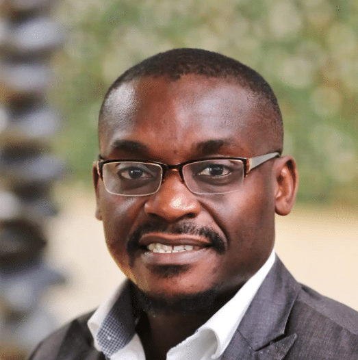 A man wearing glasses and a suit jacket smiles at the camera, with a blurred green and beige background.