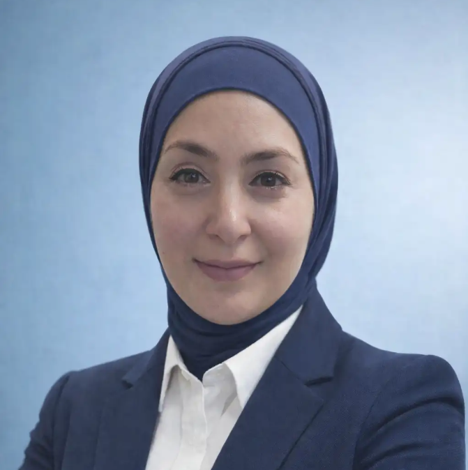 A woman wearing a navy blue hijab and suit jacket over a white shirt, posing in front of a light blue background.