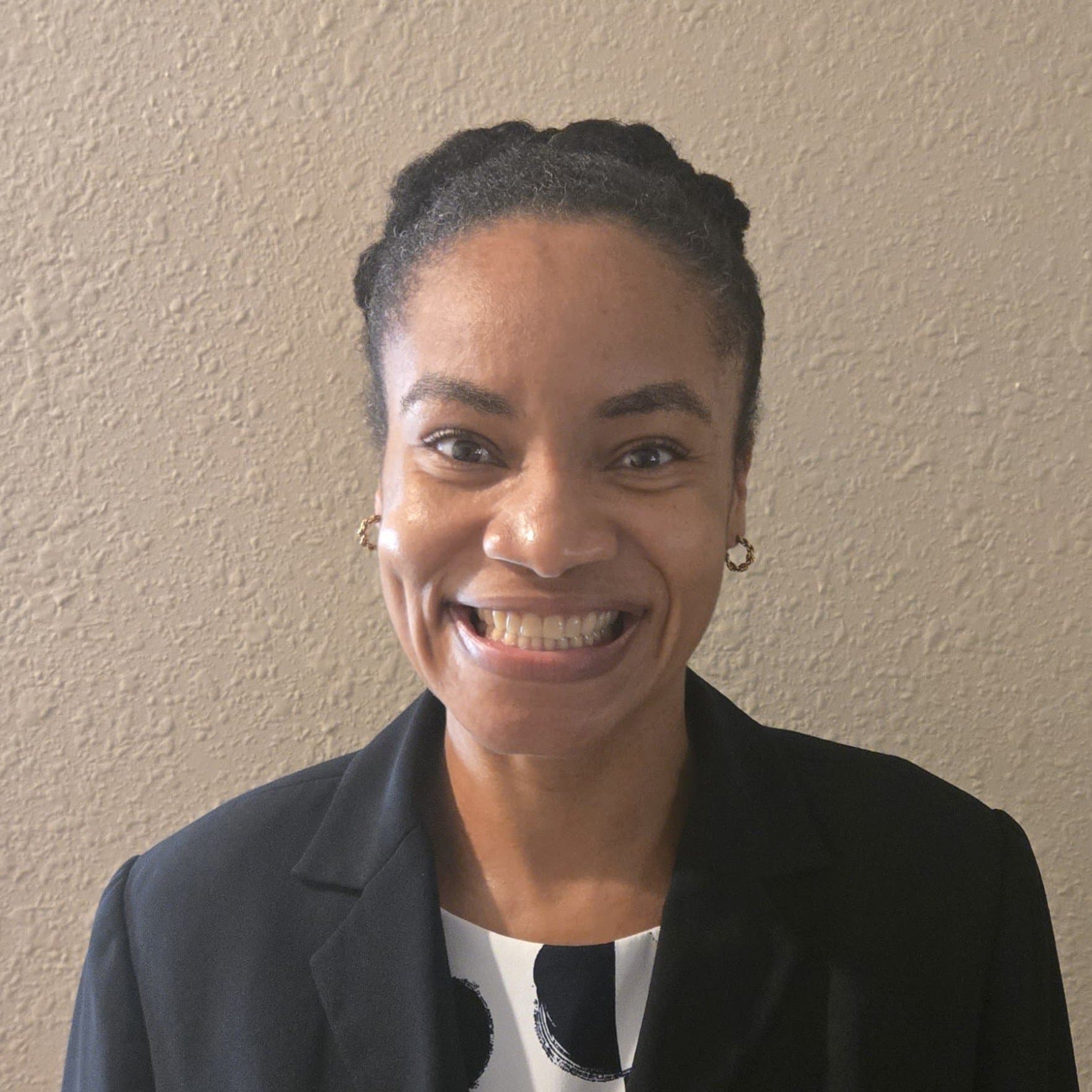 Woman with braided hair smiles at the camera, wearing a black blazer and a patterned top, standing in front of a textured beige wall.